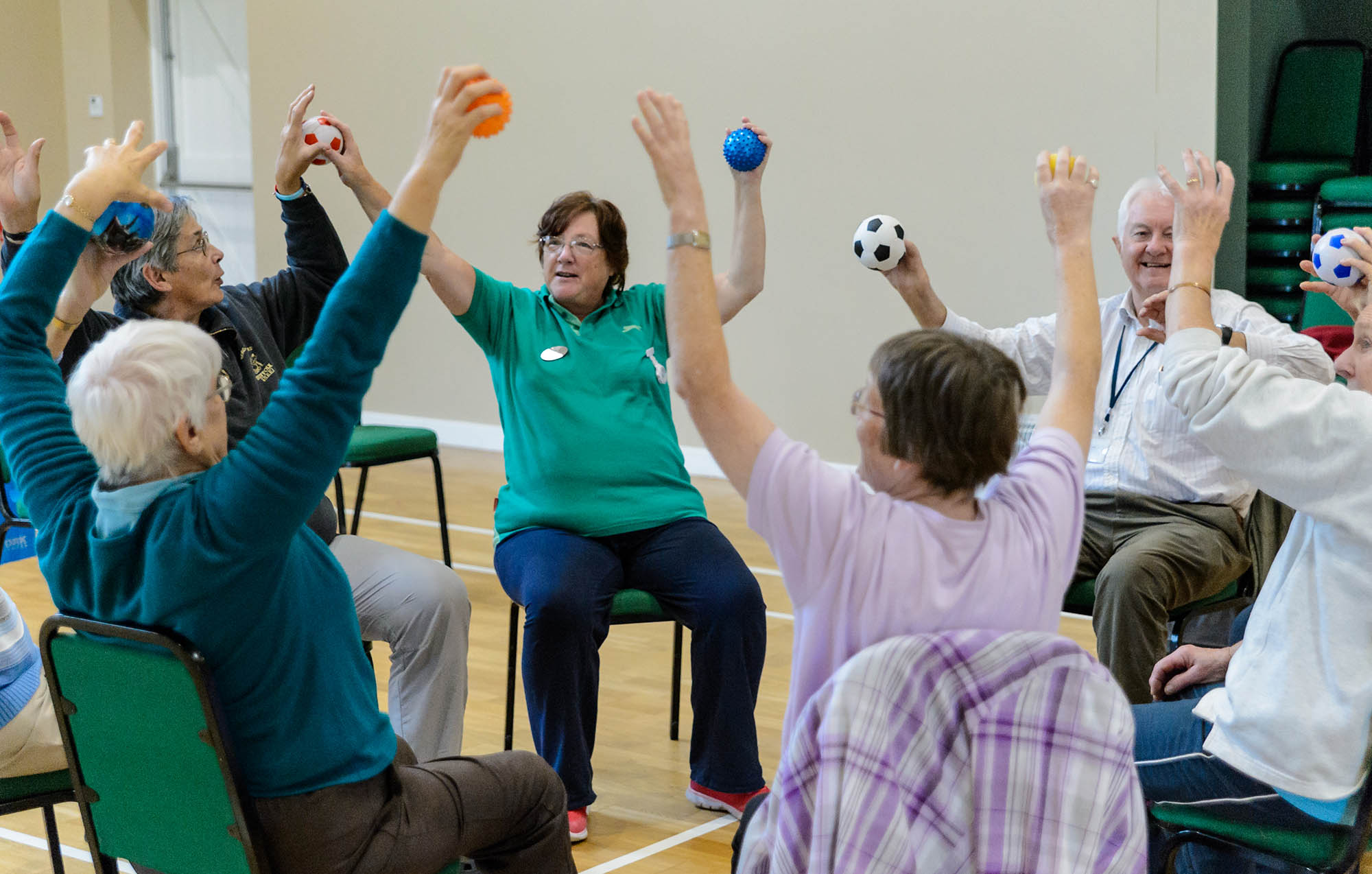 A group of older people sitting on chairs in a circle, raising their hands.