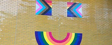 A yellow tiled building wall with a smiling mouth rainbow flag, with two eyes formed from Progress Flags. The bullding is the Manchester Metropolitan Students Union