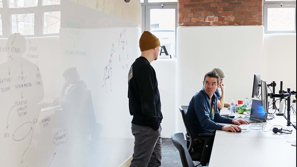 Three colleagues discussing things in an office. One is standing, two are sitting down using comptuers
