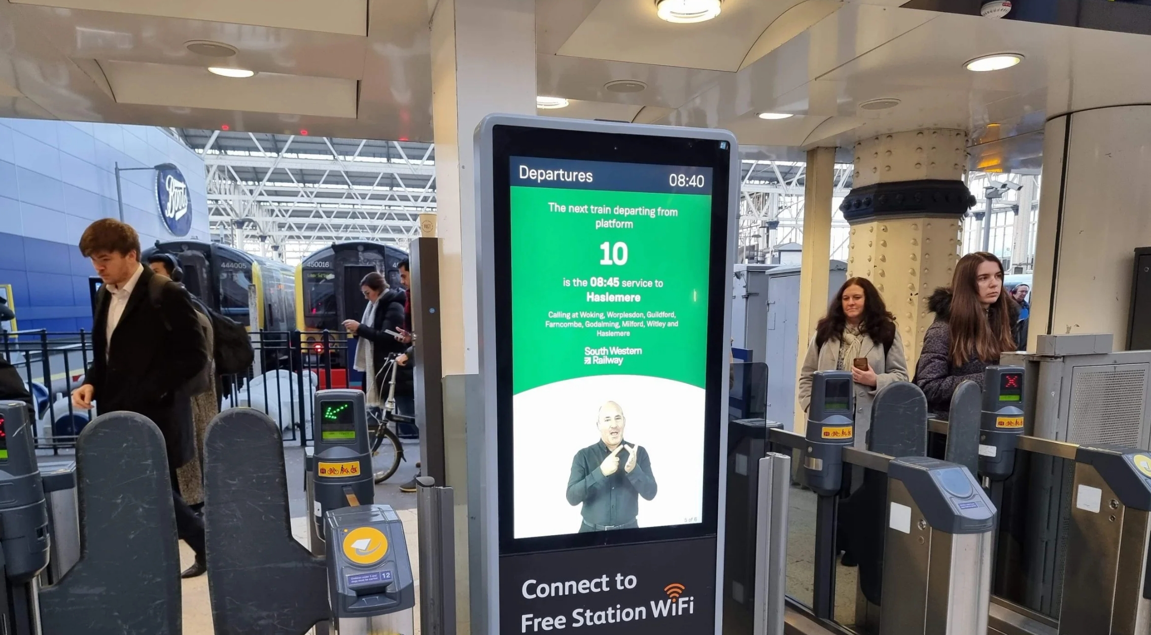 A screen shows an AI sign language tool in operation at a ticket gate, the AI generated person on screen is signing with corresponding text above