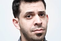 A white man with black hair and a stubbly beard. He's looking discerningly at the camera.