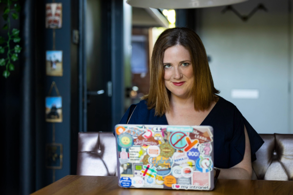 Sharon O'Dea sits at a desk. The back of her laptop is visible, and covered in stickers. She has short brown hair and blue eyes.