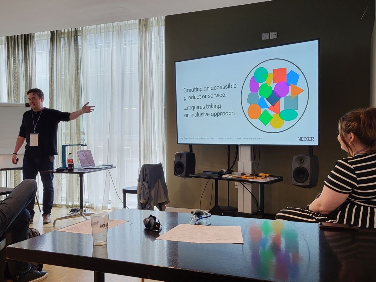 Chris Bush gestures towards a screen with a slide reading "Creating an accessible product or service...requires taking an inclusive approach"