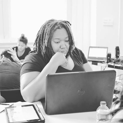 A black and white photo of a black woman with dreadlocks, looking glum and staring at a laptop.