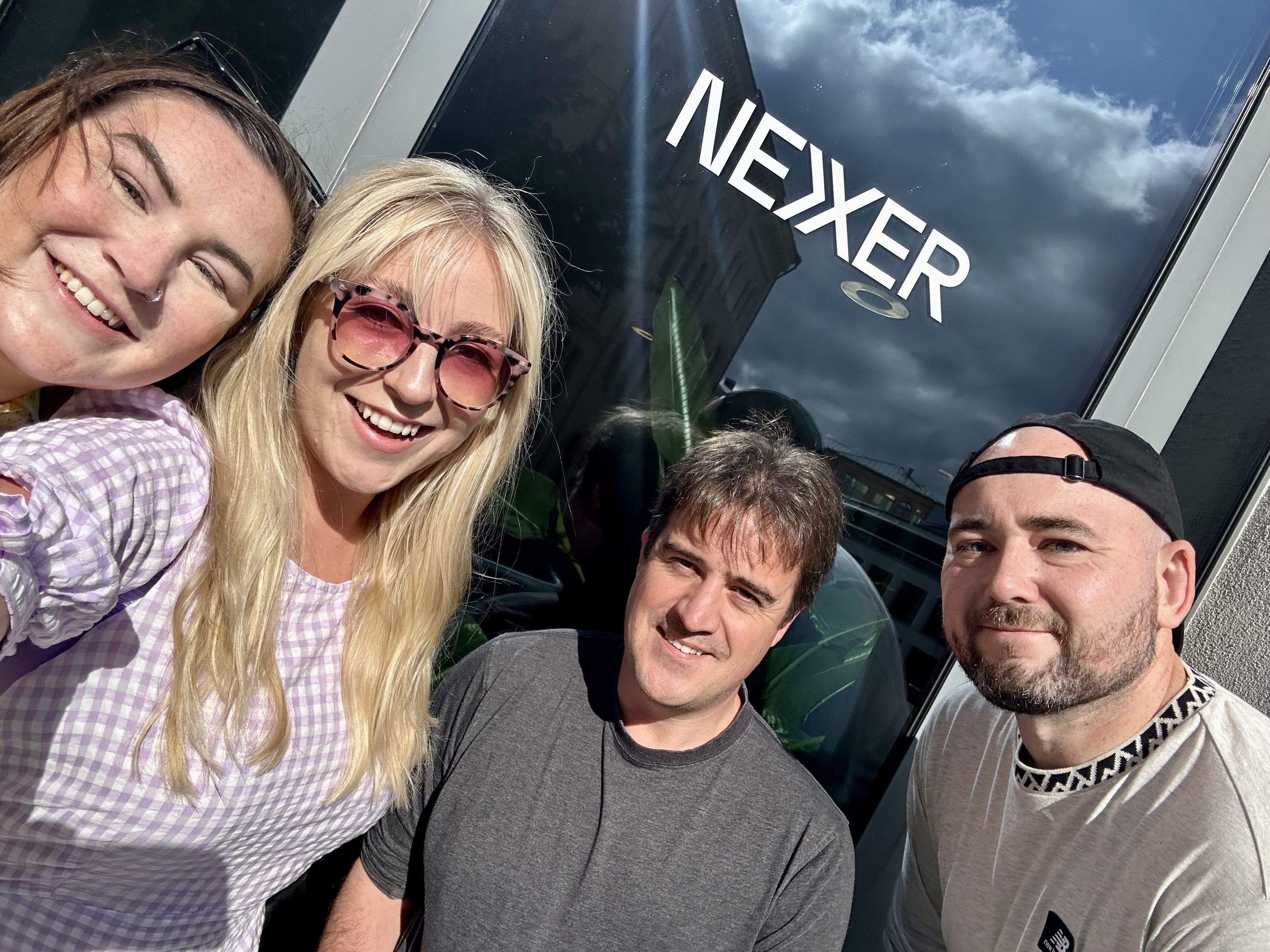 Molly, Amy, Chris and Shaun smile at the camera, taking a selfie in front of a Nexer logo
