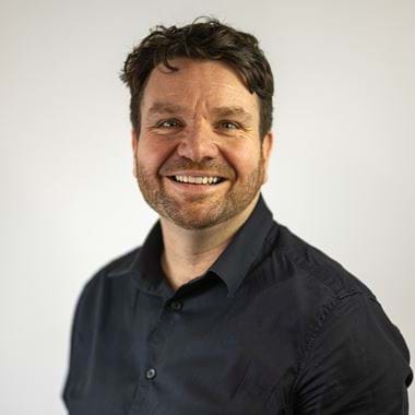 Hugh is standing against a plain white background and smiling. He has short dark hair and a beard, wearing a dark button-up shirt