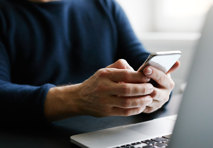 A pair of hands holding a mobile phone next to an open laptop. 
