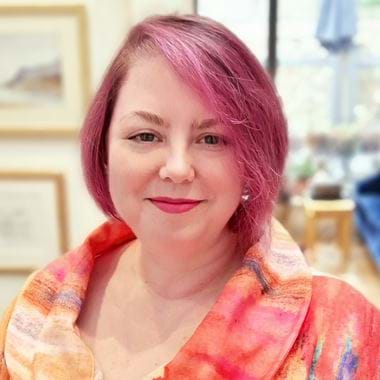 Claire is smiling at the camera. Her hair is a deep pink colour, and she is wearing a pink and orange shirt with a watercolour-esque design.