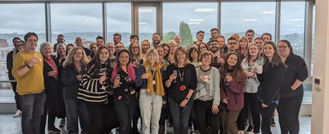 A group of about 65 people standing in an office with green hills in the background.