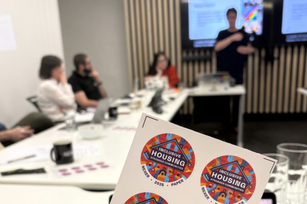 A close us shot of the stickers from the event reading "inclusive housing, stronger communities", with people visible seated in the background