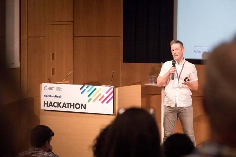 A picture of Francis Rowland, a ginger haired man with a white shirt and grey chinos, presenting at the BioData Hackathon.