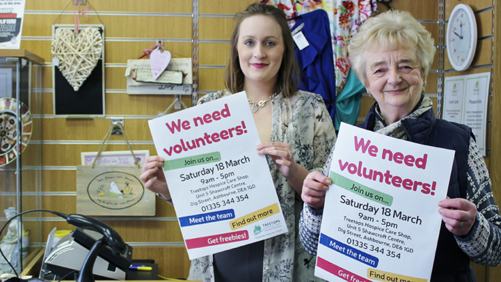 Two white women volunteering in a charity shop, holding up posters reading "We need volunteers"