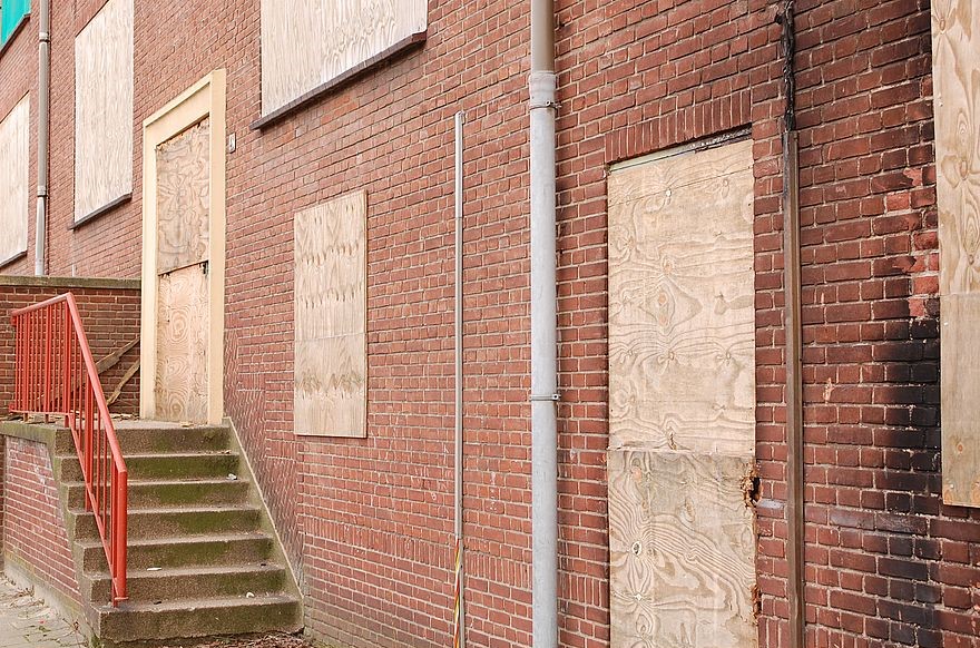 An old red brick building, with stone steps and a red railing at the front. All the windows and doors are boarded up