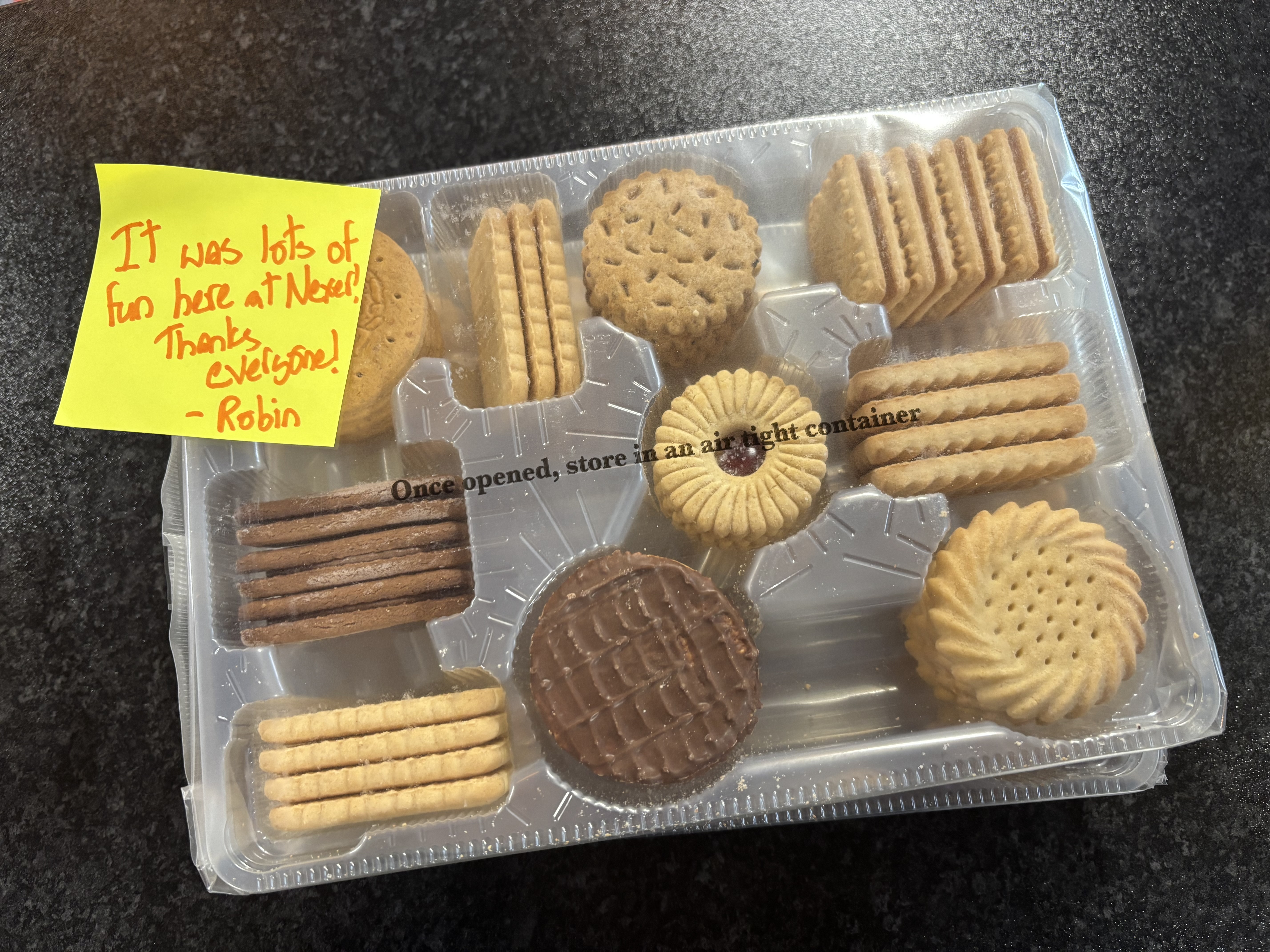 A clear plastic tray with a variety of biscuits in. A yellow post-it note is stuck to the top left corner and reads "It was lots of fun here at Nexer! Thanks everyone! Robin"