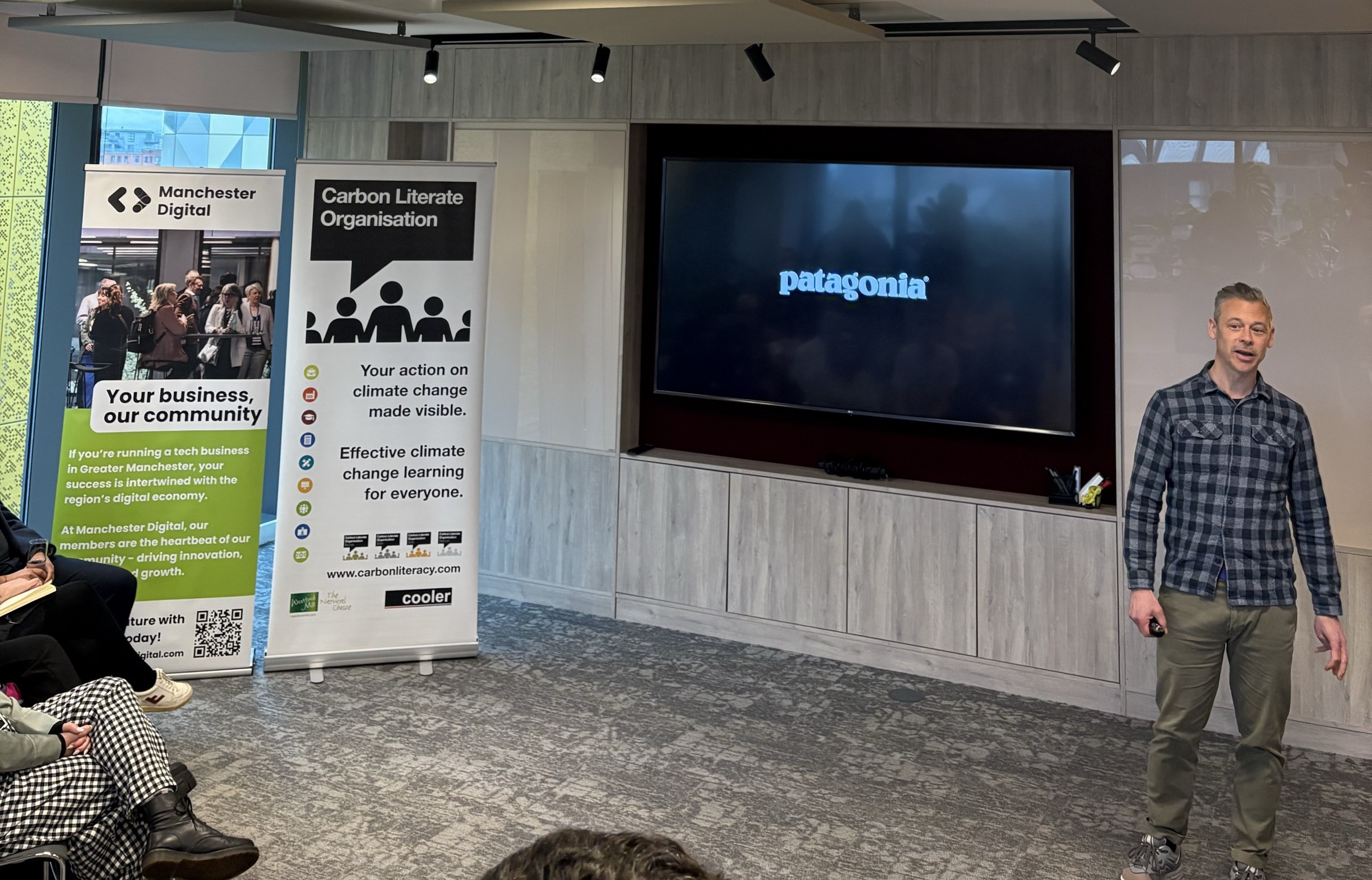 A speaker stands in front of a room of seated people. Beside him is a Carbon Literacy Toolkit rollup banner, and onscreen behind him is the logo for the brand "Patagonia"