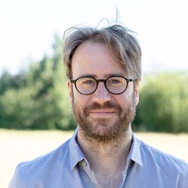 Richard is wearing glasses, has a beard, and stands outdoors, wearing a light blue shirt He is standing in an open area surrounded by trees and a on a sunny day.