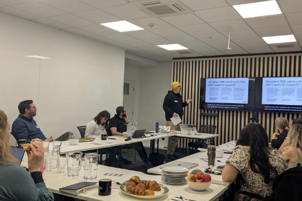 Dig Falby stands in front of a room of seated people who are taking notes and typing on laptops. He is gesturing towards a screen, featuring text about how LLMs can support users to access grants.