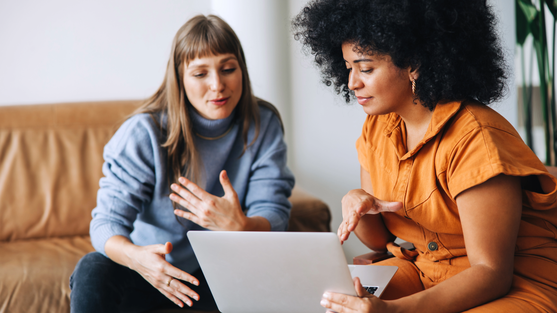 Two women sit together on a sofa, looking at a laptop and speaking