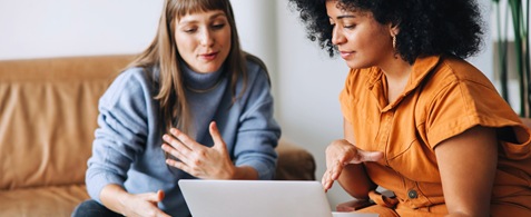 Two women sit together on a sofa, looking at a laptop and speaking