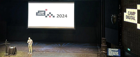 Camp Digital host Shaun Gomm stands onstage at the RNCM, with the Camp Digital logo visible behind and on the wall