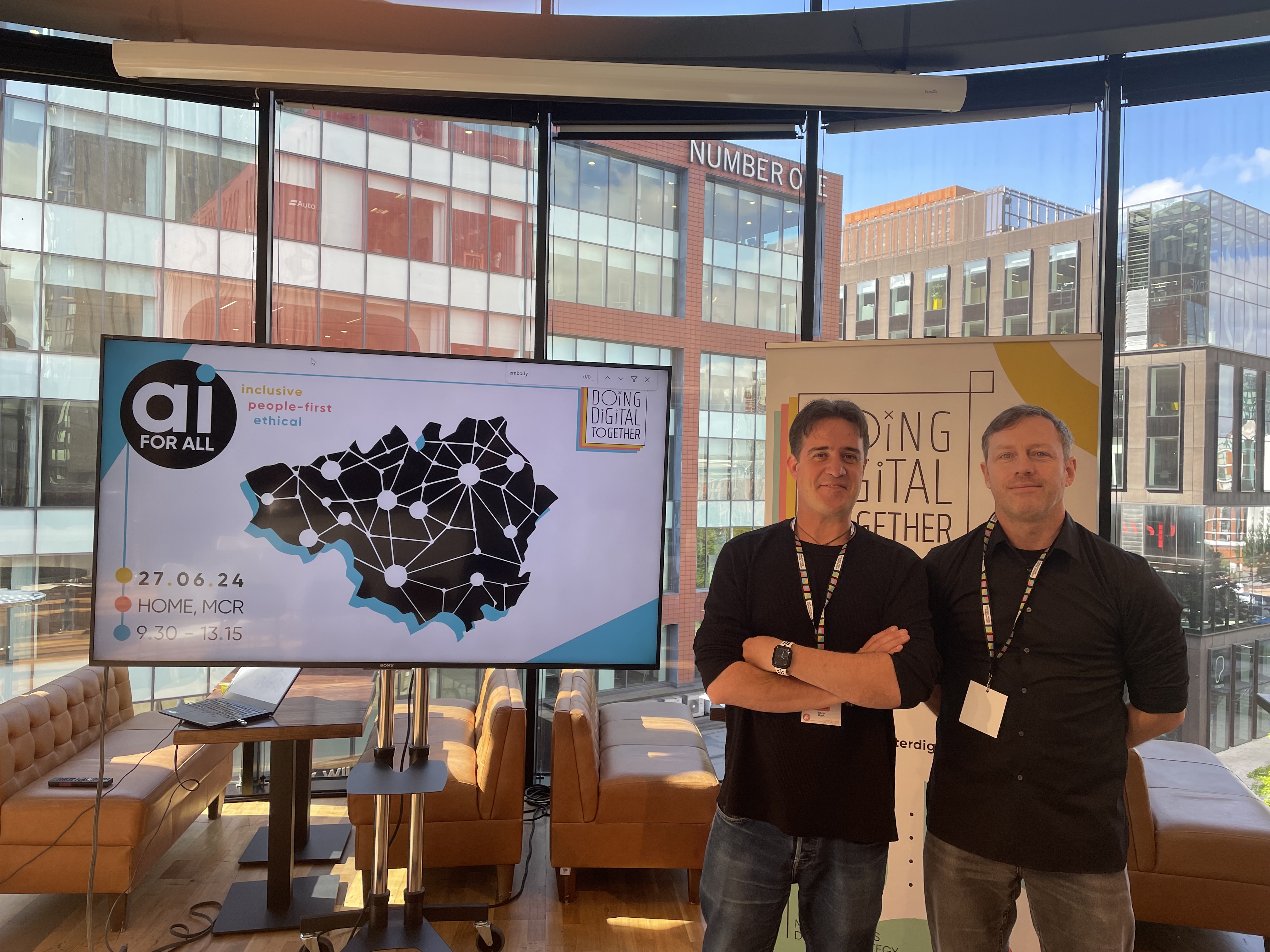 Chris and Lee are both smiling. Chris is wearing a black top and black jeans, and has brown hair and eyes. Lee is wearing a black shirt and jeans and has blue eyes and brown hair. They are both wearing lanyards, and beside them a screen reads "AI for All - People-first, inclusive, ethical"