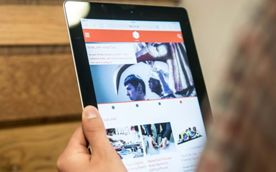 A tablet held up against a wooden wall, with the FACT website displaying on the screen. 