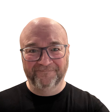 Matt has a shaved head and a beard, is wearing glasses and a black shirt, and smiling broadly at the camera. The background is blurred
