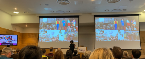 A person standing in front of two screens showing a selection of images. People are seated theatre-style in a conference hall in front of them 