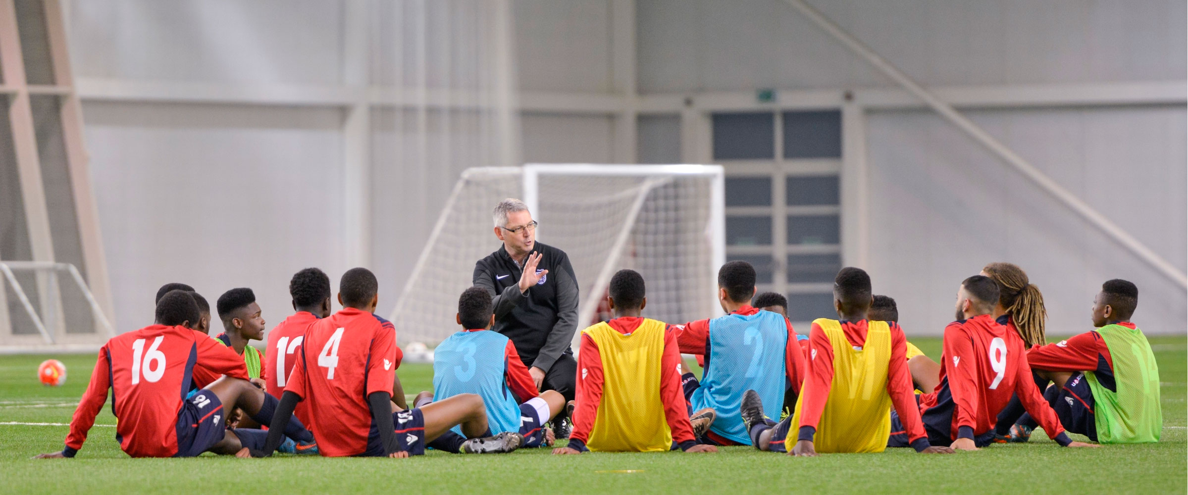 A group of football players sat on the pitch listening to their coach