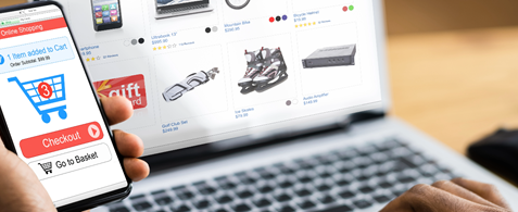 A person holds a phone in one hand, with a checkout basket displayed on the screen. On a laptop screen, being typed by the other hand, is an eCommerce interface