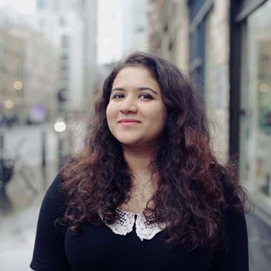 Hera grins at the camera. She has long, curly hair. Stands on a city street, wearing a black top with a white lace collar. The background is blurred, showing buildings and parked bicycles on a rainy day