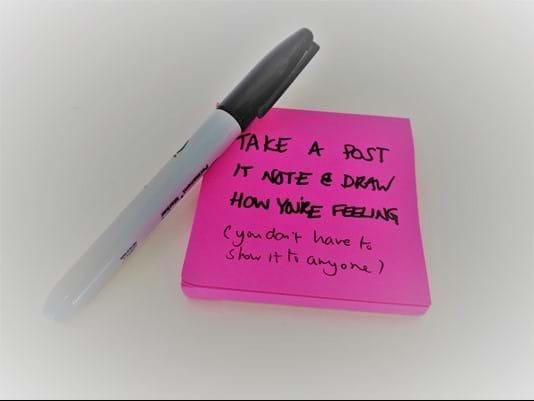 A Sharpie pen laid across the corner of a stack of pink post-its. Writing reads "Take a post it note & draw how you are feeling (you don't have to show anyone)"