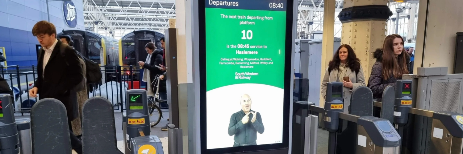A screen shows an AI sign language tool in operation at a ticket gate, the AI generated persononscreen is signing with corresponding text above