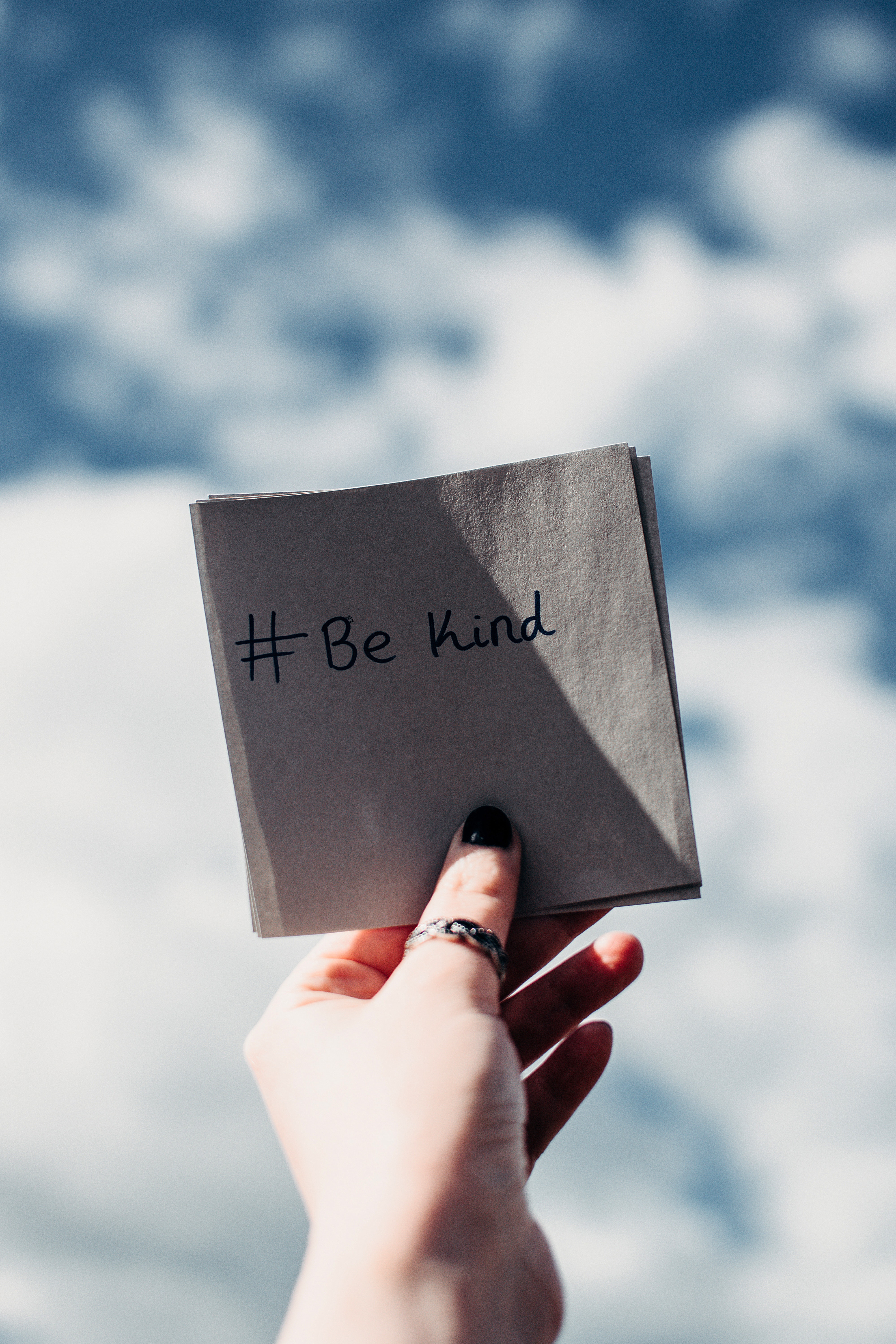 A left hand holding a note with "# Be Kind" written on it up to the sky