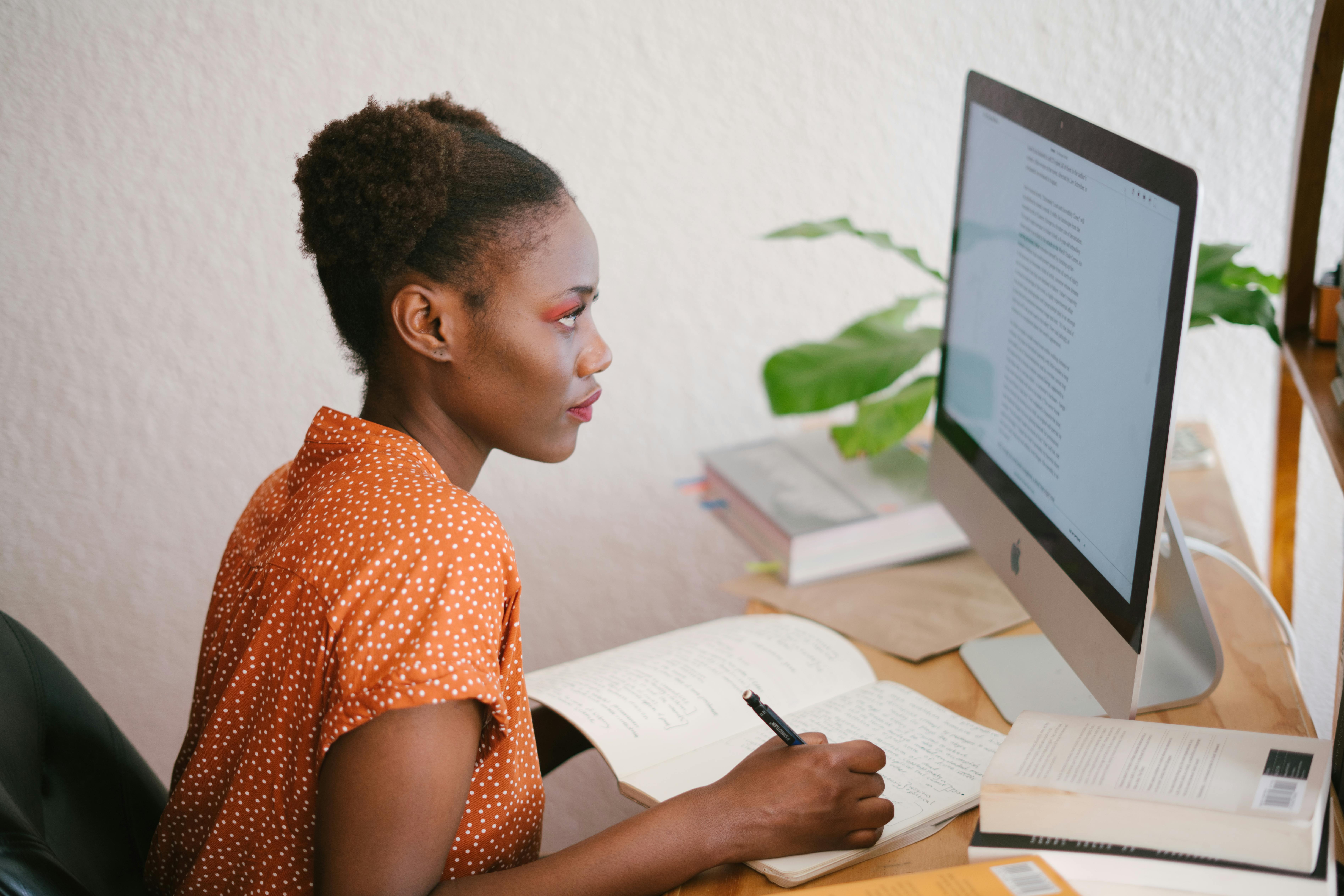 A woman is looking at a laptop screen and making notes in a notepad