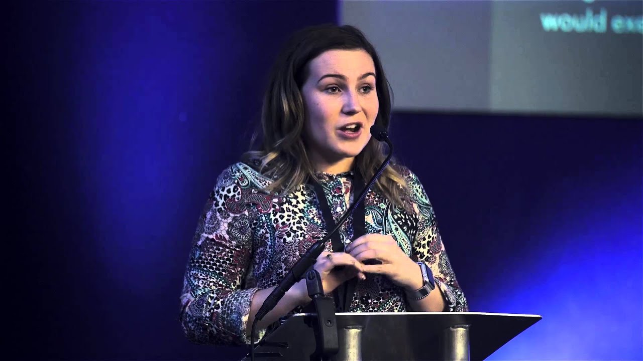 Molly Watt. A white woman with long brown hair, wearing a printed top, speaking into a microphone and standing at a podium. She is stood against a blue background. 