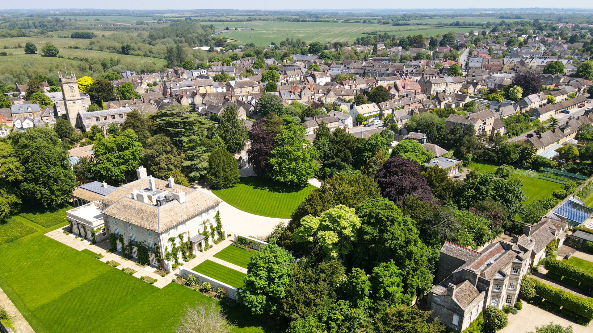 An aerial shot of Blenheim Palace, with the grand green gardens surrounding it. Beyond that houses can be seen stretching into the distance.