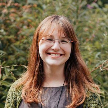 Clara is smiling in garden in Autumn. She has reddish-brown hair, wearing glasses, and a green sweater over a grey top