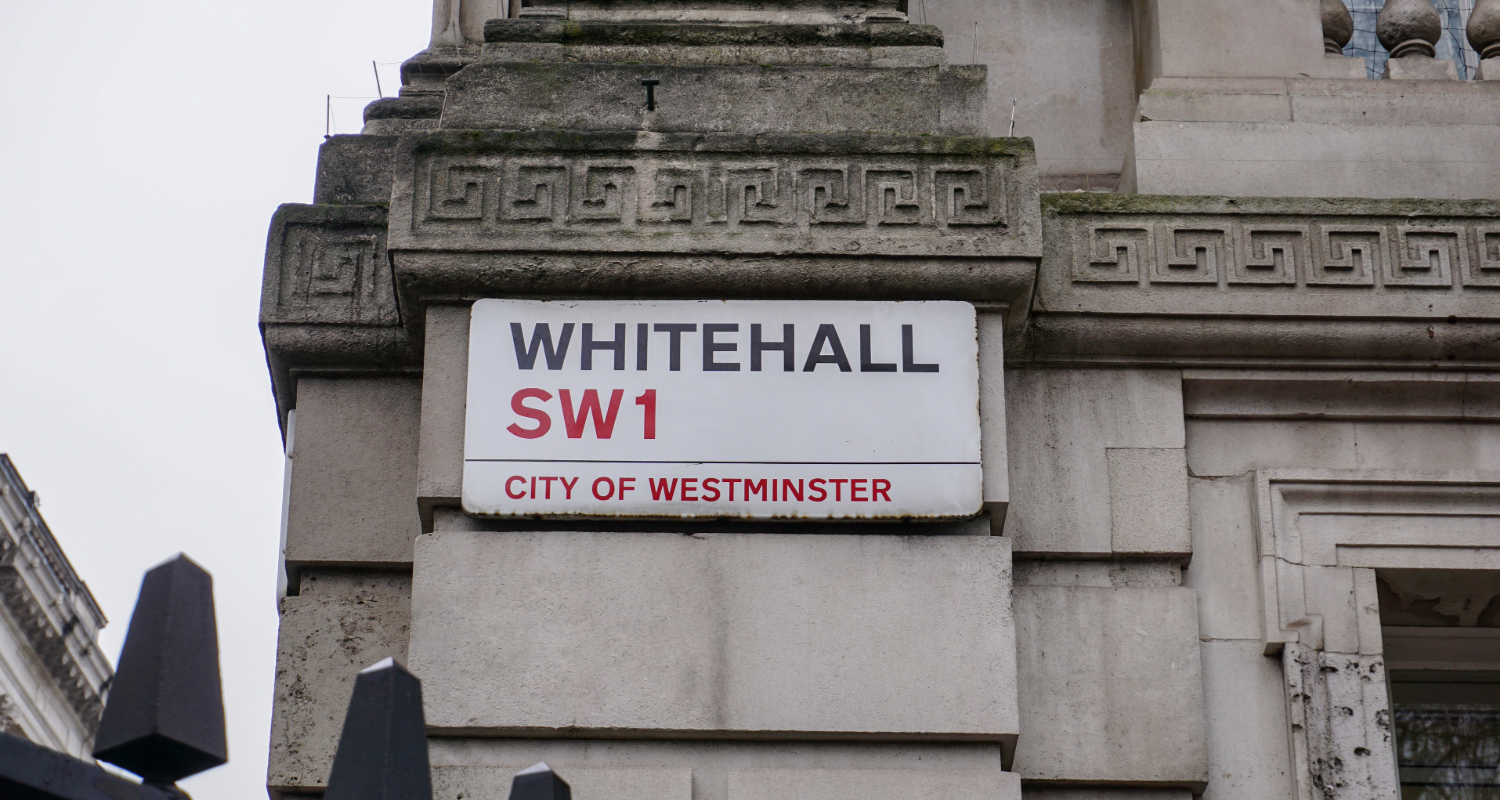A sign reading "Whitehall SW1 is mounted on a white brick building, with the top of large gates seen at the bottom of the image