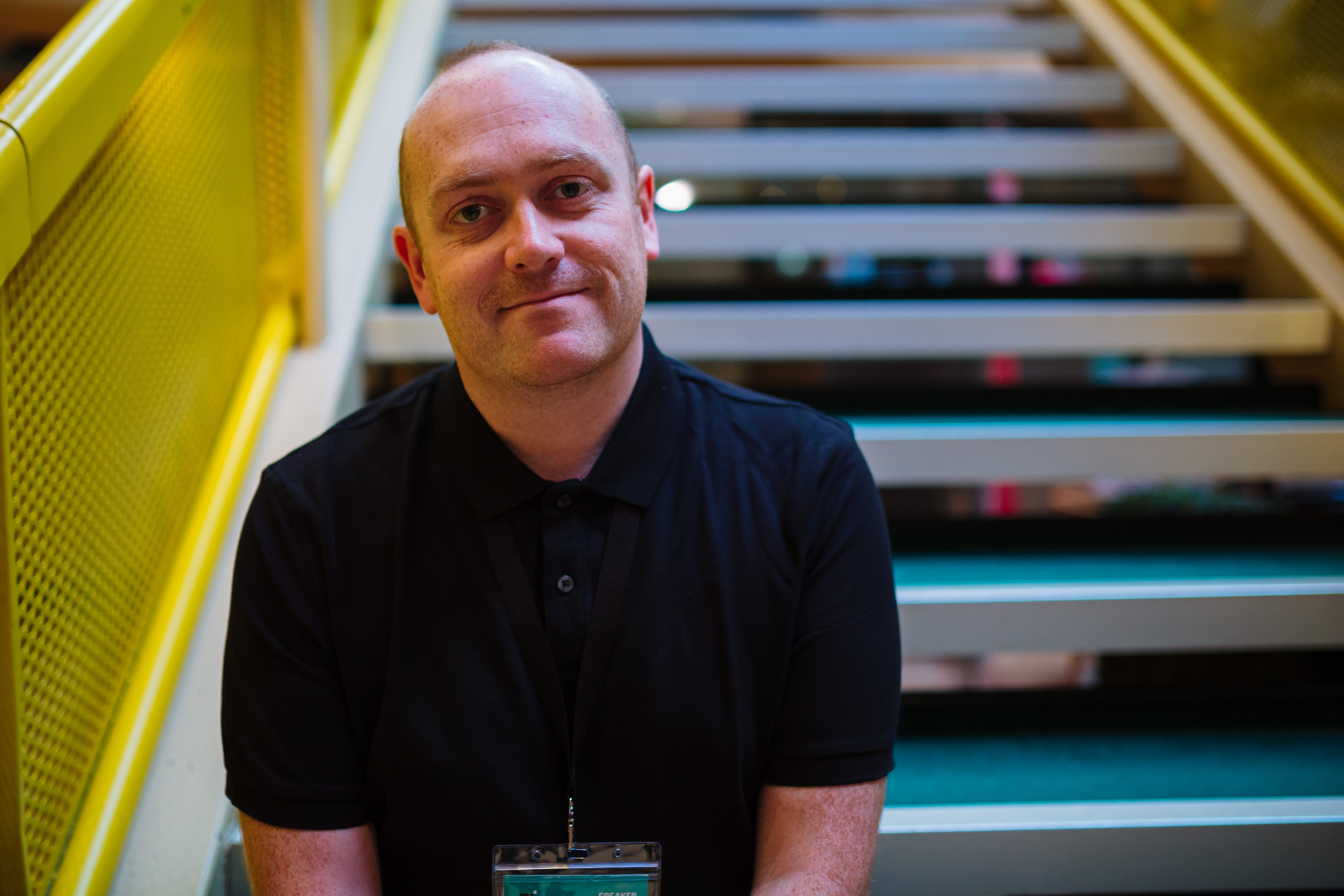 A smiling bald white man in a black t-shirt. He is sat on stairs with a yellow bannister. 